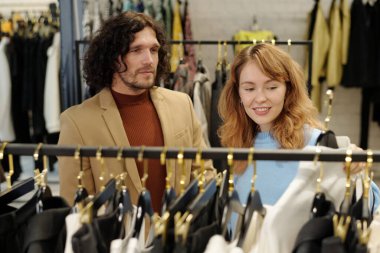 Mutlu genç kadın ve kocası şık giyinmiş ve butikte yeni moda koleksiyonuna bakıyorlar.