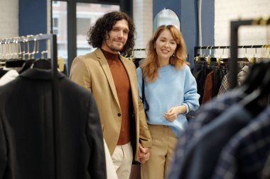 Sessiz, lüks giysiler içinde genç ve sevecen bir çift butikte duruyor ve yeni sezonluk moda kıyafetlere bakıyor.
