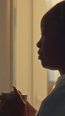 Nöbete gitmeden önce pencerenin önünde durup kahve içip iş hakkında düşünen melankolik Afro-Amerikan sağlık görevlisinin dikey çekimleri.