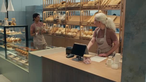 年轻的女人把新鲜的面包放在货架上 然后去收银机 帮同事在面包店干活 — 图库视频影像