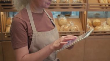 Önlüklü yaşlı, gri saçlı bir kadının ekmek raflarının yanında durup fırında dijital tablet üzerinde çalışmasını çek.