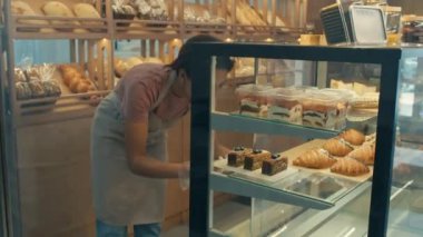 Tek kullanımlık eldivenli kadın çalışanın çekimleri fırındaki raflara tatlılar koyuyor.