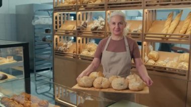 Önlüklü yaşlı kadın fırıncının portresi. Elinde bir tepsi ekmek ve fırında çalışırken kameraya gülümsüyor.