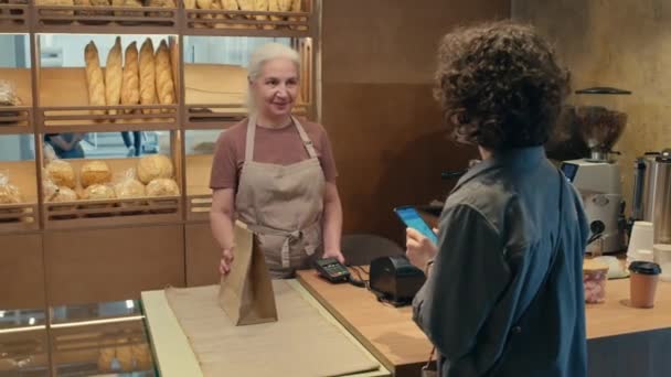 Cliente Femenino Pagando Con Smartphone Agradeciendo Vendedora Senior Llevando Bolsa — Vídeo de stock