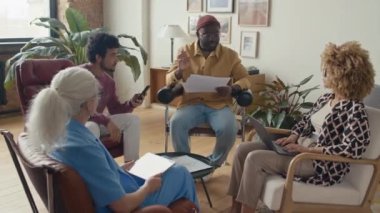 Gündelik giysiler içinde Afrika kökenli Amerikalı takım lideri gündemi modern ofiste oturan çeşitli meslektaşlarına anlatıyor.