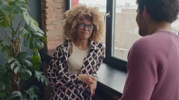 身着休闲装和眼镜的年轻非洲裔美国女商人靠在窗台上 一边与男同事聊天 一边在办公室休息 — 图库视频影像