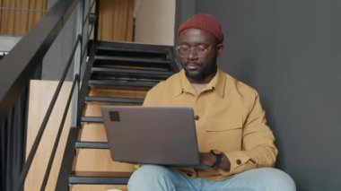 Rahat ofisteki merdivenlerde oturan, dizüstü bilgisayar kullanan ve kameraya gülümseyerek poz veren gündelik kıyafetli genç Afrikalı Amerikalı adamın portresi.