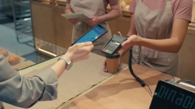 Kadın fırın işçisinin terminali tutarken, müşterinin akıllı telefonla para ödemesini ve kahve almak için alışveriş yapmasını çek.