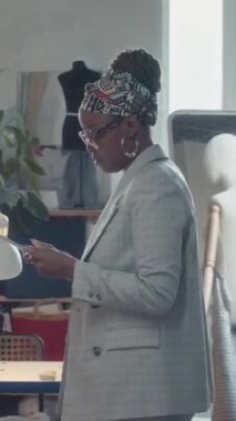 Gözlüklü ve tabletli Afrikalı Amerikalı kadın atölye patronunun dikey hâli. Kıyafet üretim sürecinde dikiş makineleri üzerinde çalışan çeşitli terzileri kontrol ediyor.