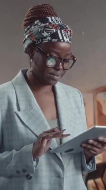 Gri takım elbiseli ve gözlüklü, tablet kullanan ve arka planda çalışan terzilerle kameraya bakan şık gülümseyen Afrikalı Amerikalı kadının dikey portresi.