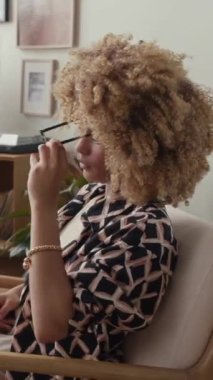 Gen Z 'nin dikey portresi kıvırcık saçlı, gözlük takan, kameraya poz veren ve ofiste çalışırken gülümseyen Afro-Amerikan iş kadını.