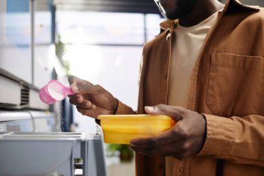 Yıkama tozu kepiyle genç Afrikalı Amerikalı adamın yakın plan fotoğrafı. Otomatik makineyle özel bölüme dökülüyor.