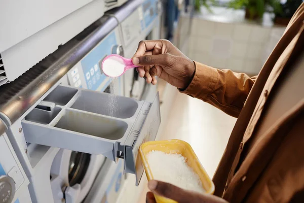 Hand of African American guy in beige shirt putting cap of soap powder into special grey plastic compartment of washing machine