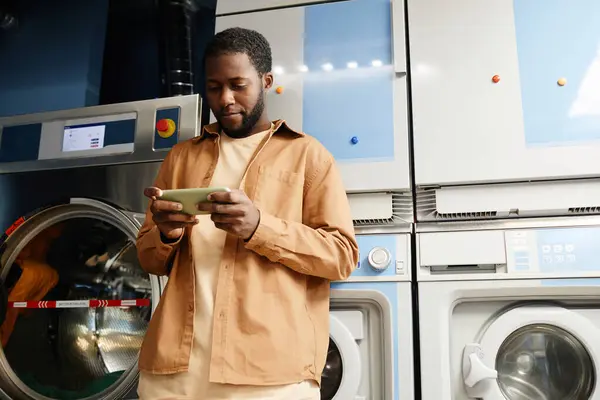 Young African American man marking time of washing in his smartphone or watching online video while standing by automatic machine