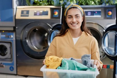 Giysiler içinde güzel bir kız temiz giysilerle plastik bir kap tutuyor çamaşırhanede çamaşır makinelerine karşı ve kameraya bakıyor.