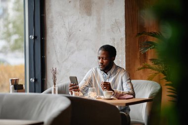 Ciddi bir Afrikalı beyaz yakalı işçi kafede masada otururken cep telefonu ekranına bakıyor ve çerezlerle kahve içiyor.