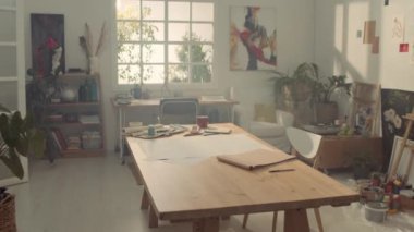 Büyük ahşap masa, kağıt, boya ve diğer malzemeler, duvarlarda resimler ve eskizler, güneşli bir sabahın erken saatlerinde, sanatçılar veya ressamlar stüdyosunun iç çekimleri.