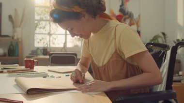 Stüdyoda tekerlekli sandalyede oturan, resim defterine boya kalemleriyle resim çizen, resim dersine katılan engelli, gülümseyen genç kadın sanatçı mercekleriyle orta boy fotoğraf.