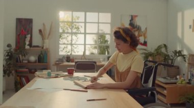 Orta halli, engelli genç beyaz kadın ressamın atölyede tekerlekli sandalyede oturup pastelle resim çizerken ve neşeyle gülümserken, güneşli bir sabahta