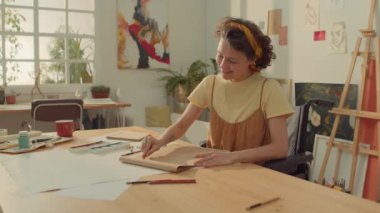 Stüdyoda tekerlekli sandalyede oturan, eskiz defterine pastel boyalarla resim çizen ve güneşli bir günde tek başına çalışırken gülümseyen orta boylu, beyaz kadın ressam.