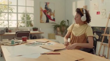 Sanat stüdyosunda tekerlekli sandalyede oturan, pastel boyayla resim çizen, kulaklıkla müzik dinleyen, ritimle sallanan ve şarkı söyleyen orta halli genç kadın sanatçı.