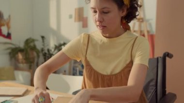 Orta ölçekli, engelli, tekerlekli sandalyede oturan, atölye masasında oturan, eskiz defterine pastel boyalarla soyut çizgileri ve şekiller çizen beyaz kadın.