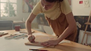 Orta halli beyaz kadın sanatçı stüdyoda masada durmuş, hevesle kağıt üzerine pastel tebeşirle resim çiziyor, sonra da kontrol ediyor.