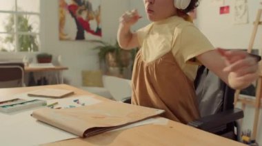 Boyama atölyesinde tekerlekli sandalyede oturan, Pastel 'le resim çizen, kulaklıkla enerjik müzik dinleyen ve sallanan engelli genç kadının orta boy yakın çekimi.