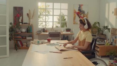 Sanat stüdyosunda tekerlekli sandalyede oturan, pastel boyayla resim çizen, kulaklıkla müzik dinleyen, ritimle sallanan ve şarkı söyleyen orta halli genç kadın sanatçı.