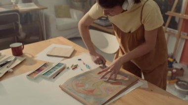 Orta açılı, kulaklıklı neşeli beyaz kadın sanatçının atölyede pastelle soyut bir resim çizerken, favori müziği dinlerken, birlikte şarkı söylerken, zıplarken ve ritme göre sallanırken.