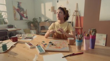 Orta boy melez kadın resim öğretmeni atölyede oturuyor, eskiz kitabı, pastel resim, fırça ve boya ile, ve sosyal medya içeriğini çekerken resim dersine başlıyor.