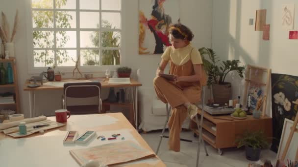 全景照片 忙碌的白人女艺术家身穿连衣裙和T恤衫坐在高椅子上 一边在工作室里一边用铅笔在笔记本上画草图 一边看着窗户 — 图库视频影像