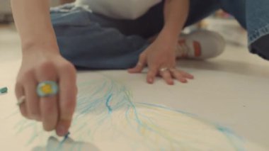 İsimsiz kadın ressamın ya da kot pantolonlu kızın elleri, parmaklarında yüzükler, atölyede yerde oturuyor, resim çiziyor, çizimleri mavi tebeşirle çiziyor.