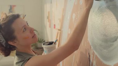 Kafkasyalı neşeli, kıvırcık saçlı, atölyede soyut resim yapan, elinde siyah boya kovası tutan ve sünger ve parmaklarıyla dairesel şekiller çizen beyaz kadının yakın çekimi.