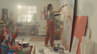 Tulum giymiş kadın dekoratörün stüdyoda elinde boya tepsisiyle dikilirken ve geniş bir çalı çırpı tabakasına soyut çizgiler çizerken tam bir yan görüntüsü.