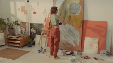 Tulum giymiş, kıvırcık saçlı, atölyedeki ekstra geniş ve soyut sanat eserleri üzerinde çalışan, kontrplak çarşafa sarı boya sıçratan isimsiz kadın ressamın tam arkası.