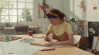 Orta ölçekli, siyah kıvırcık saçlı, sanatçı atölyesinde masa başında oturan beyaz bir kadın resmi ve gelecekteki yaratıcı başyapıt için kahverengi çizim defterine kurşun kalemle bir şeyler çiziyor.