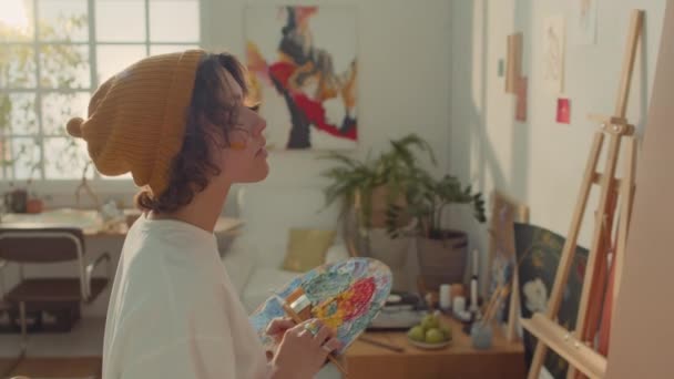 身披便帽 心事重重的年轻女画家身临其境 站在画架前 手握彩色调色板 构思未来艺术品的设计 — 图库视频影像