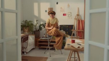 Geniş açıda, tulumlu beyaz kadın ressam evdeki rahat sanat stüdyosunda yüksek taburede oturuyor, çizim defterine kurşun kalemle çiziyor ve pencereye bakıyor.