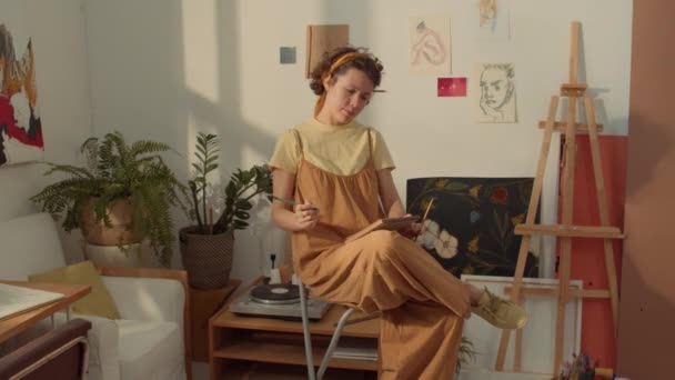 カーリーヘアの若い白人女性のミディアムフルショット アートスタジオのハイスツールに座ってジャンプスーツ 鉛筆でスケッチブックを描いた後 ペンシルベニアフェイスで窓を見下ろす — ストック動画