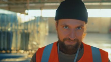 Kafkasyalı gülümseyen sakallı depo çalışanının güvenlik önlüğü giymiş ve iş günü kameraya bakan siyah bereli portresi.