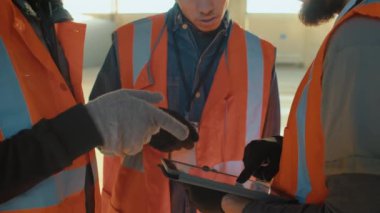 Orta bölüm, üç inşaat işçisinden oluşan, güvenlik giysisi ve pamuk eldivenli, kapalı tablet ekrana bakan fikirleri tartışan tanımlanamayan bir ekibin görüntüsü.