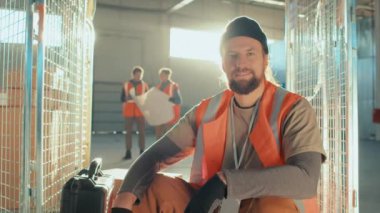 Gülümseyen beyaz işçinin orta boy portresi. Alet çantasıyla depodaki kameraya bakıyor.