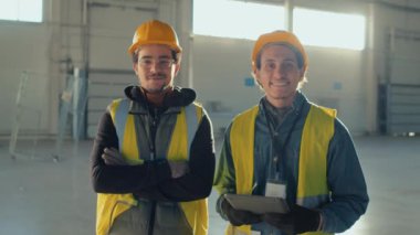 İki çift ırklı inşaat işçisinin orta boy portresi. Kasketli ve koruyucu iş kıyafetli. Gülümseyen ve kameraya tabletle poz veren.