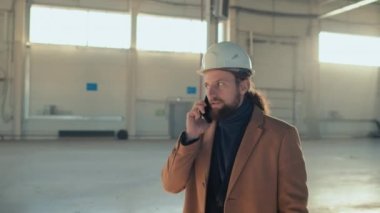 Beyaz güvenlik kasketi, takım elbise ve bej ceket giyen beyaz sakallı bina sahibinin kapalı alanda inşaat alanında yürürken akıllı telefondan konuşmasının görüntüleri.