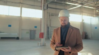 Beyaz sakallı bir adamın tabletle ve proje tasarım uygulaması planlamasıyla iç inşaat sahasında yürürken orta boy görüntüsü.