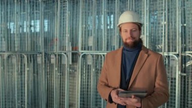 Beyaz kasketli beyaz sakallı mühendisin orta boy portresi ve depodaki kameraya bakarak, tabletle depoda poz veren günlük kıyafetler.