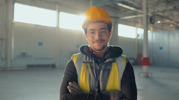 阳光明媚的一天 在空旷的室内建筑工地 拍摄年轻戴着眼镜 头戴安全帽 头戴双手交叉相机的年轻建造者的肖像 — 图库视频影像