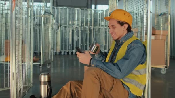 在仓库休息期间 戴着安全帽 头戴安全帽 头戴手机坐在购物车上 用金属杯子喝茶或喝咖啡的年轻男性员工的侧影 — 图库视频影像