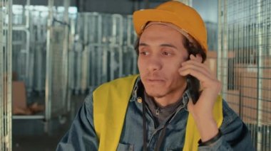 Sırıtkan, turuncu kasklı ve güvenlik üniformalı melez işçinin depo molasında akıllı telefondan konuşma görüntüsü.